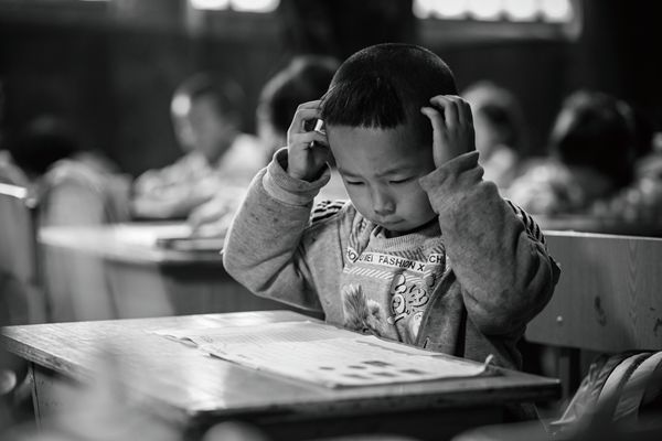 A young Chinese boy scratching his head as he sits at his desk reading a book. 